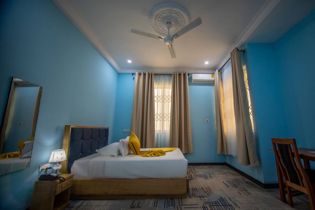 Une chambre d’hôtel avec un lit, une table de chevet, une lampe, un ventilateur de plafond et une fenêtre avec des rideaux.