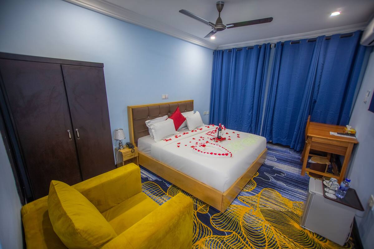 Une chambre d’hôtel avec un lit, un fauteuil jaune, une armoire, un bureau avec une lampe et un ventilateur de plafond.
