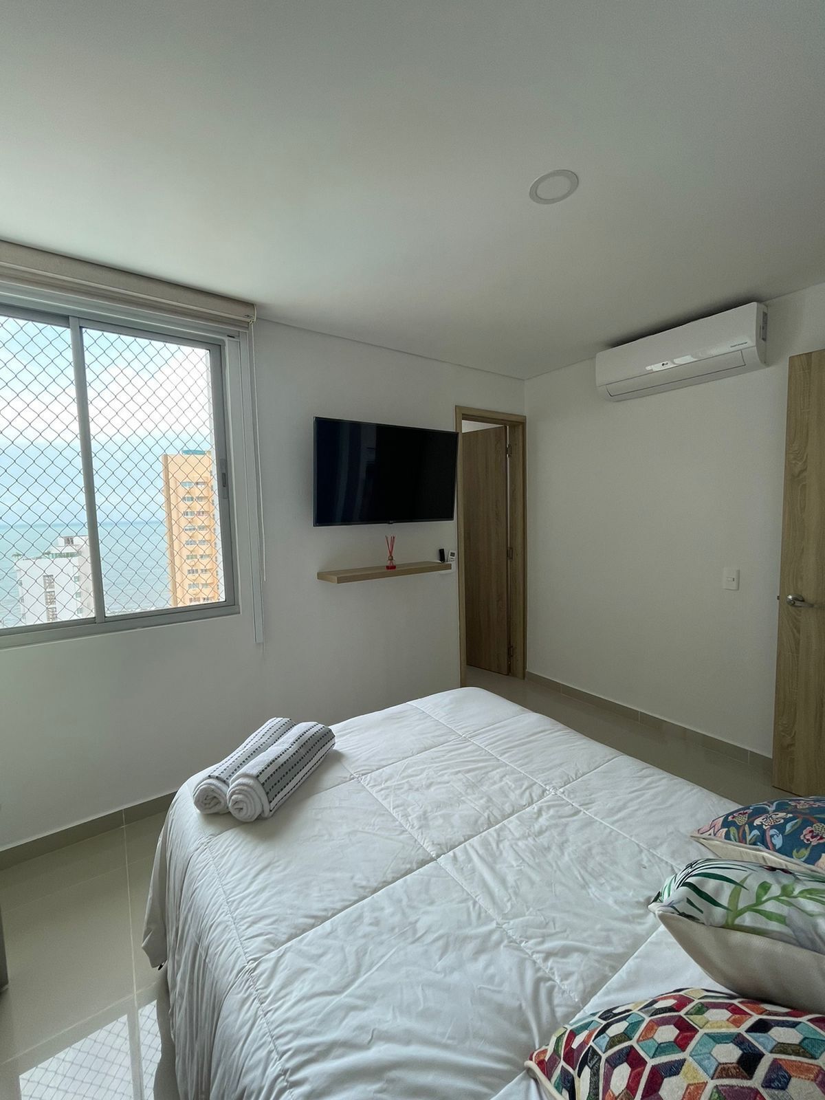 A bedroom with a bed, a TV mounted on the wall, an air conditioning unit, and a window with a view of buildings.