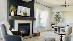 Uma sala de estar com lareira, televisão fixada em uma prateleira de madeira, área de refeições com mesa e cadeiras, e uma grande janela com cortinas.