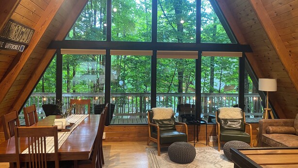 A spacious room with a large wooden table, several chairs, a sofa, and a large window offering a view of a forest. - Apres’ Haus: Comfy A-Frame centrally located in Whitecap Village with fireplace  (Upson)