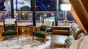 A cozy, well-lit room with a large window overlooking a snowy landscape. There are two leather sofas and two armchairs with fluffy cushions. - Apres’ Haus: Comfy A-Frame centrally located in Whitecap Village with fireplace  (Upson)