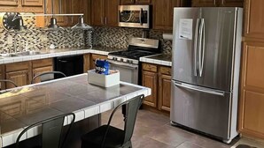 A kitchen with wooden cabinets, a stainless steel refrigerator, a microwave, and a gas stove. There is a bar stool and a small island with a bag of chips.
