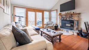 A spacious living room with a large sofa, a fireplace, a television mounted on the wall, and large windows with blinds. - Your Gateway to Pocono! (Tannersville)