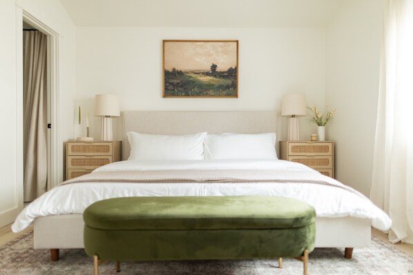 Une chambre à coucher avec un grand lit, deux tables de chevet avec des lampes, un banc vert et un tableau représentant un paysage, encadré, accroché au mur.