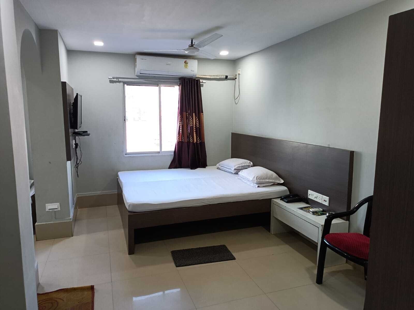 A hotel room with a bed, a desk with a chair, an air conditioning unit, a window with curtains, and a television.