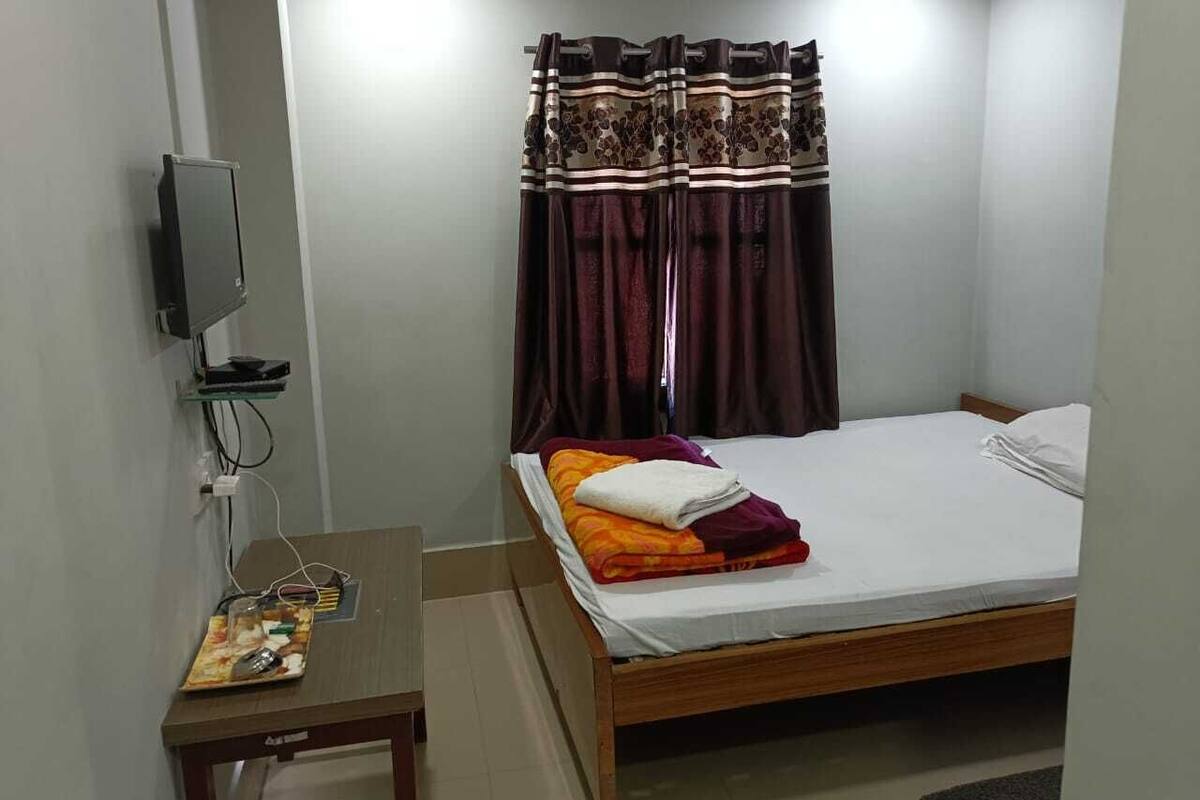Un dormitorio pequeño y ordenado con una cama individual, una mesita de noche, un televisor montado en la pared y una ventana con cortinas.