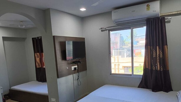 A hotel room with two beds, a TV mounted on the wall, an air conditioning unit, and a window with curtains. - Kusum Hotel And Restaurant (Dibrugarh)