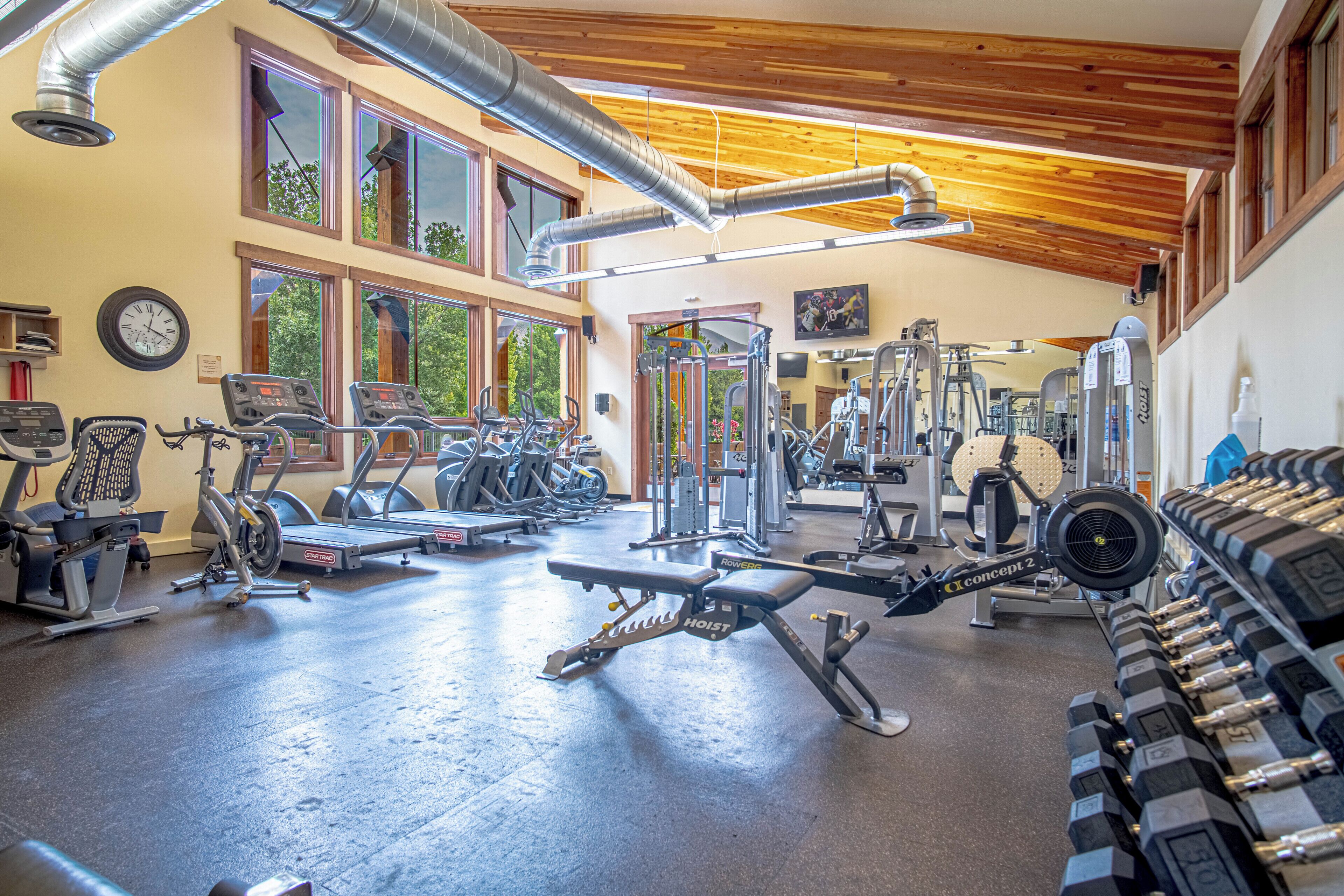 Une salle de sport équipée de divers appareils d’exercice, dotée de grandes fenêtres et d’un plafond en bois.