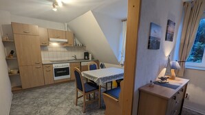 A compact kitchen with a dining area, a small table, and chairs. There is a window with curtains, a lamp on a side table, and a shelf with kitchen utensils. - Haus Friesenglück, Whg. Leuchtturm - Haus Friesenglück (Alkersum)
