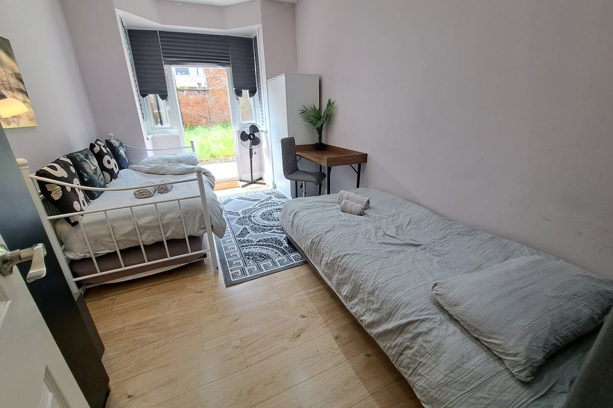 Une petite chambre bien éclairée, avec un lit, un bureau et une chaise. Une fenêtre donne sur un mur de briques et sur la verdure à l’extérieur.