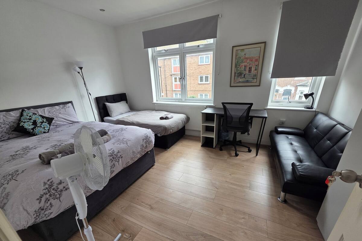 Une chambre avec deux lits, un bureau, une chaise, un canapé, un ventilateur sur pied et un radiateur.