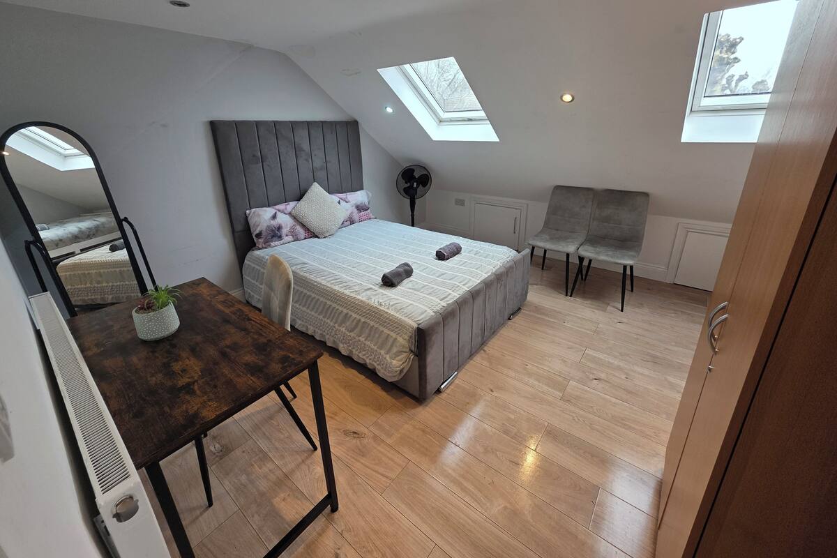 Une chambre moderne avec une lucarne, un lit doté d’une tête de lit grise, une table de chevet avec une plante et un fauteuil.