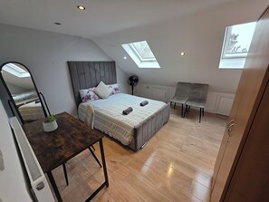 A modern bedroom with a skylight, a bed with a grey headboard, a bedside table with a plant, and a chair. - Comfy Fenia Stay (London)
