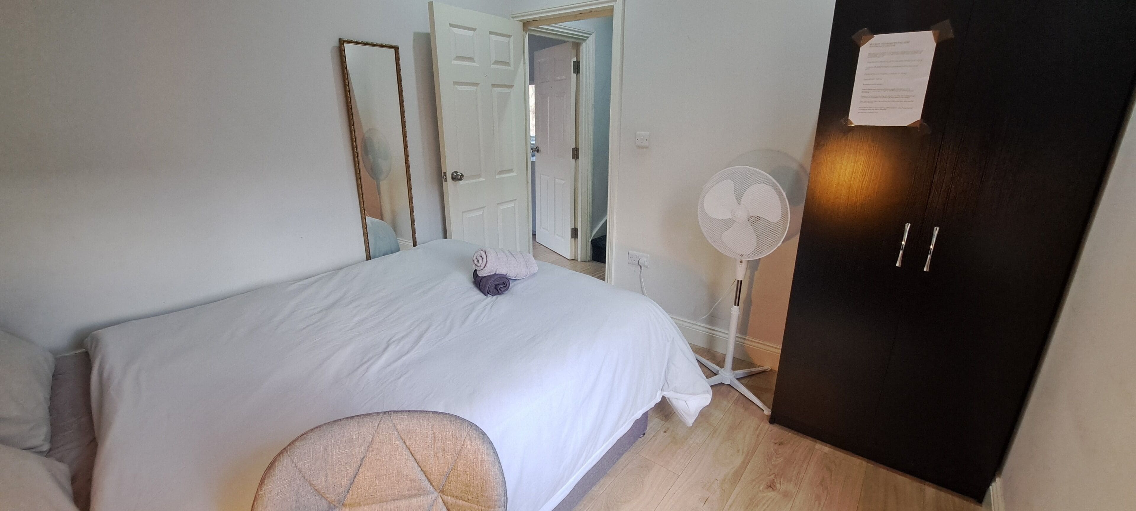 A bedroom with a bed, a bedside table, a chair, a wardrobe, a mirror, a fan, and a door.