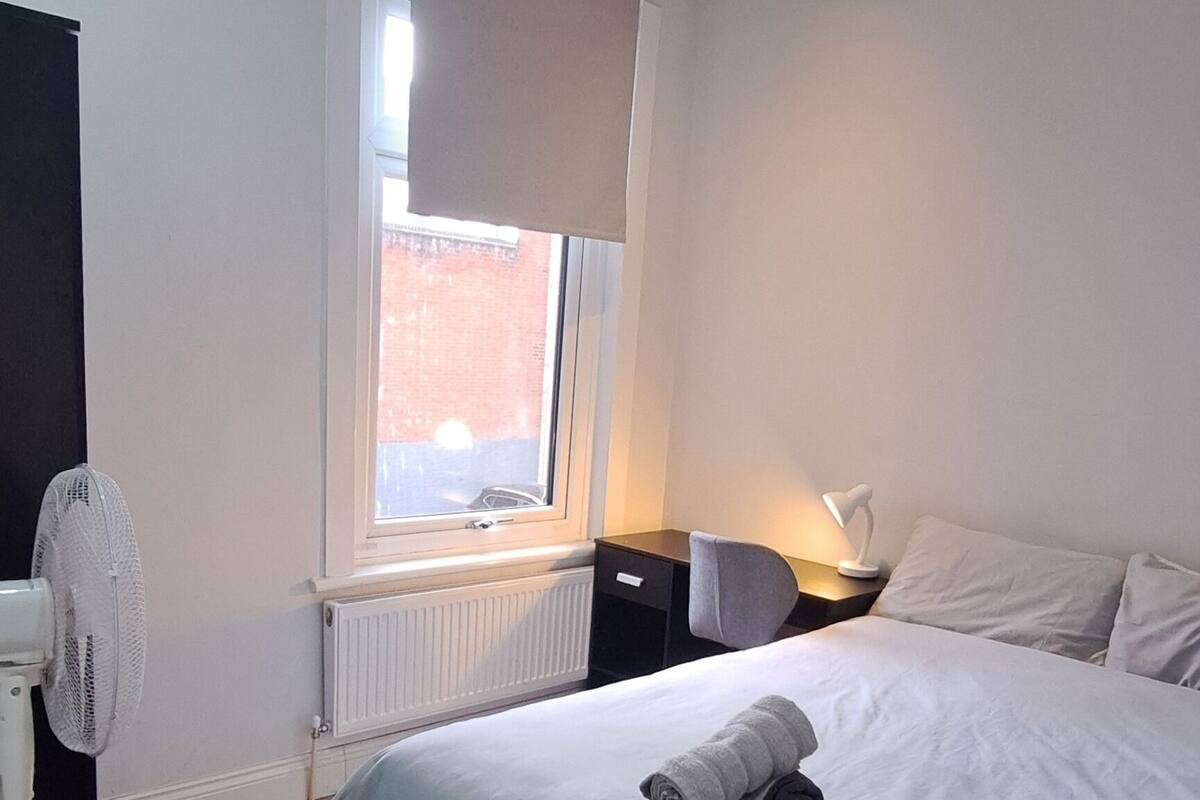 Une chambre d’hôtel moderne équipée d’un grand lit, d’un bureau, d’une chaise, d’un ventilateur sur pied et d’une fenêtre donnant sur un immeuble en briques.