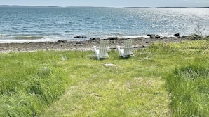A grassy area with two white Adirondack chairs facing a rocky shoreline and a calm sea under a clear sky.