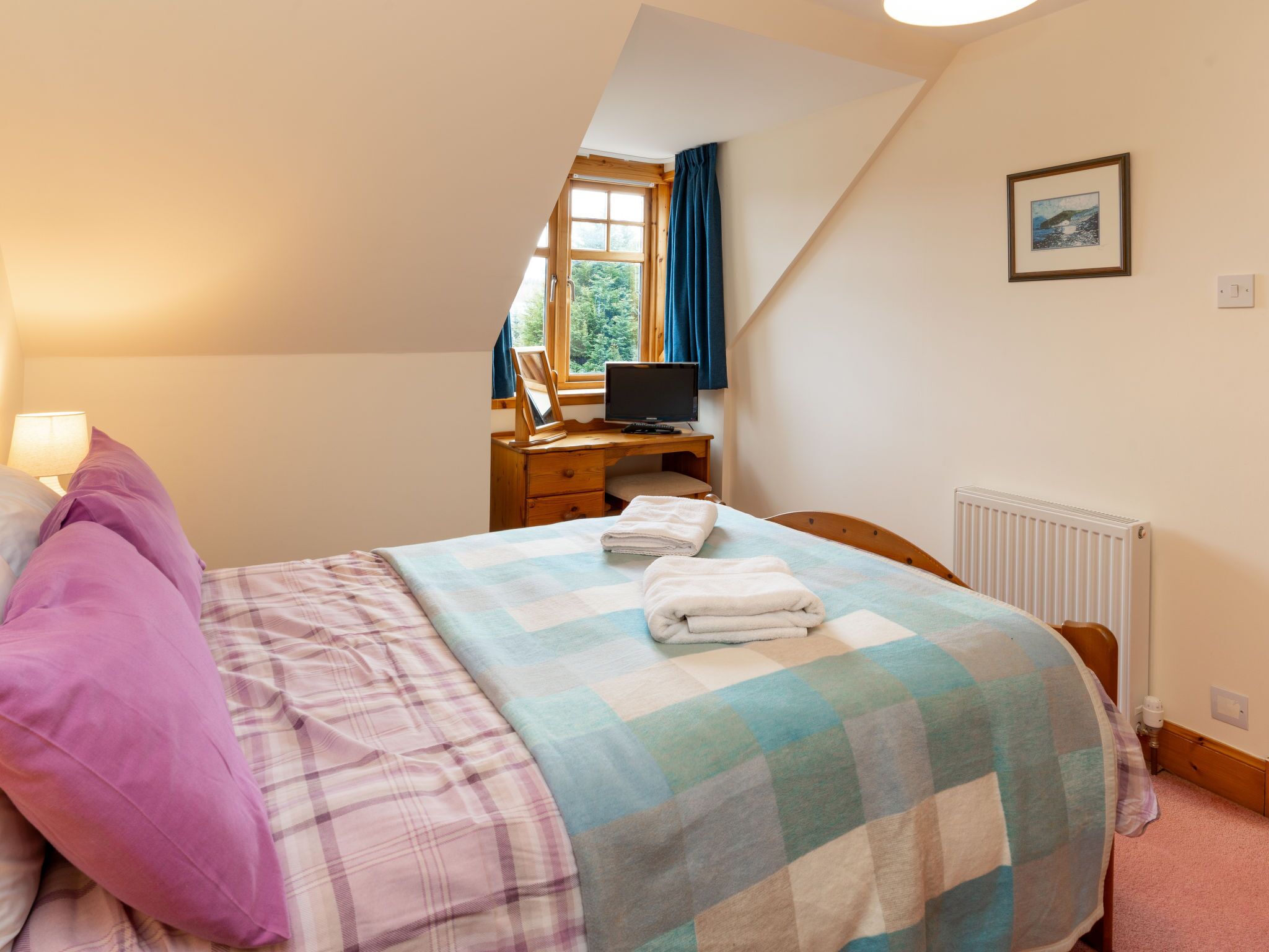 Ein Schlafzimmer mit Bett, Schreibtisch mit Computer, Fenster mit Vorhängen und Heizkörper.