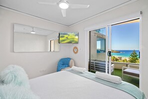 Une chambre avec un lit, un ventilateur de plafond, une télévision, une vue sur la plage et un balcon avec une balustrade.