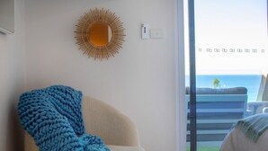A room with a large window, a chair with a blue blanket, a sunburst wall decoration, and a view of the ocean.