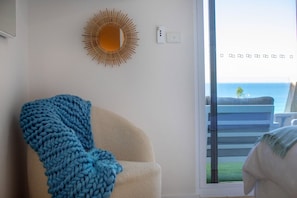 Una habitación con un ventanal grande, una silla con una manta azul, un adorno de sol en la pared y vista al océano.