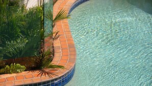 A clear swimming pool with a tiled edge, surrounded by lush green plants.