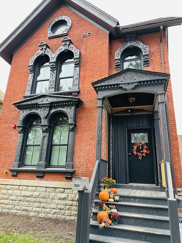 Um edifício de tijolos com detalhes em preto ornamentados, uma porta frontal preta com uma grinalda e um pequeno alpendre com abóboras e flores.