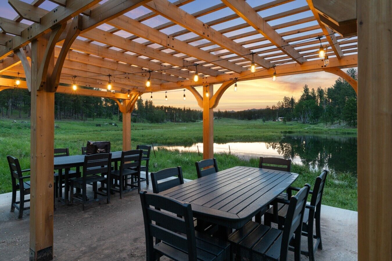 A wooden pergola with tables and chairs, overlooking a pond and forest.