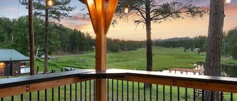 A wooden deck with a railing, a table, and chairs, overlooking a grassy area with trees and a cabin.