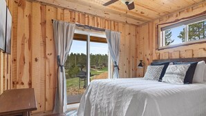 A wooden cabin bedroom with a bed, curtains, a window, and a view of the outdoors.
