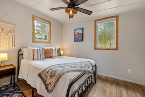 A bedroom with a bed, a ceiling fan, two windows, a nightstand, and a wall-mounted lamp.