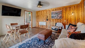 Een knusse woonkamer met een houten wand, een flatscreen televisie, een eethoek met stoelen en een bank met kussens.
