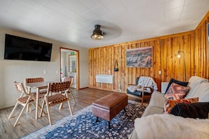 A cozy living room with a wooden wall, a flat-screen TV, a dining area with chairs, and a sofa with cushions.