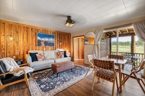 A cozy room with a wooden wall, a sofa, a coffee table, a dining table with chairs, and a view of the outdoors.