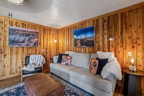 A cozy room with wooden panel walls, a grey sofa with patterned cushions, a brown ottoman, and two wall-mounted televisions displaying scenic landscapes.