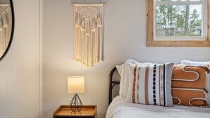 Een bed met gestreepte kussens, een bedlamp, een macramé wanddecoratie en een raam met uitzicht op bomen.