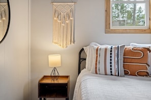 A bed with patterned pillows, a bedside lamp, a macrame wall hanging, and a window with a view of trees.