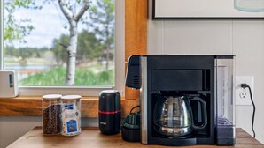 Een koffiezetapparaat, een koffiemolen en twee pakken melk staan op een houten aanrecht, vlakbij een raam met uitzicht op bomen en een wateroppervlak.