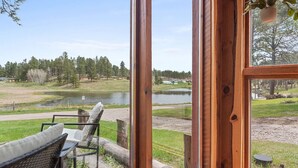 A wooden-framed window with a view of a lake, chairs, and a hanging plant.