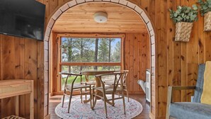 A cozy room with wooden walls, a round table with chairs, a TV, and a large window offering a view of trees.