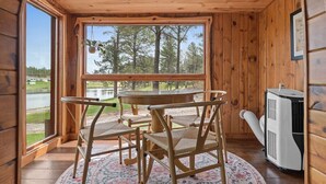 A wooden cabin interior with a round table and chairs, a large window overlooking a lake and trees, and a portable air conditioning unit.