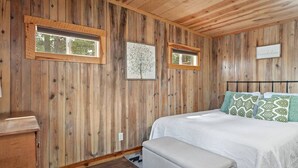A bedroom with wooden walls, a bed with patterned pillows, and two windows. - Little Joy: Needle Spire Views + Sauna + Wildlife (Custer)