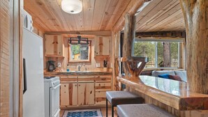 A wooden cabin interior with a kitchen, wooden walls, a window overlooking the outdoors, and a bench by the window. - Little Joy: Needle Spire Views + Sauna + Wildlife (Custer)