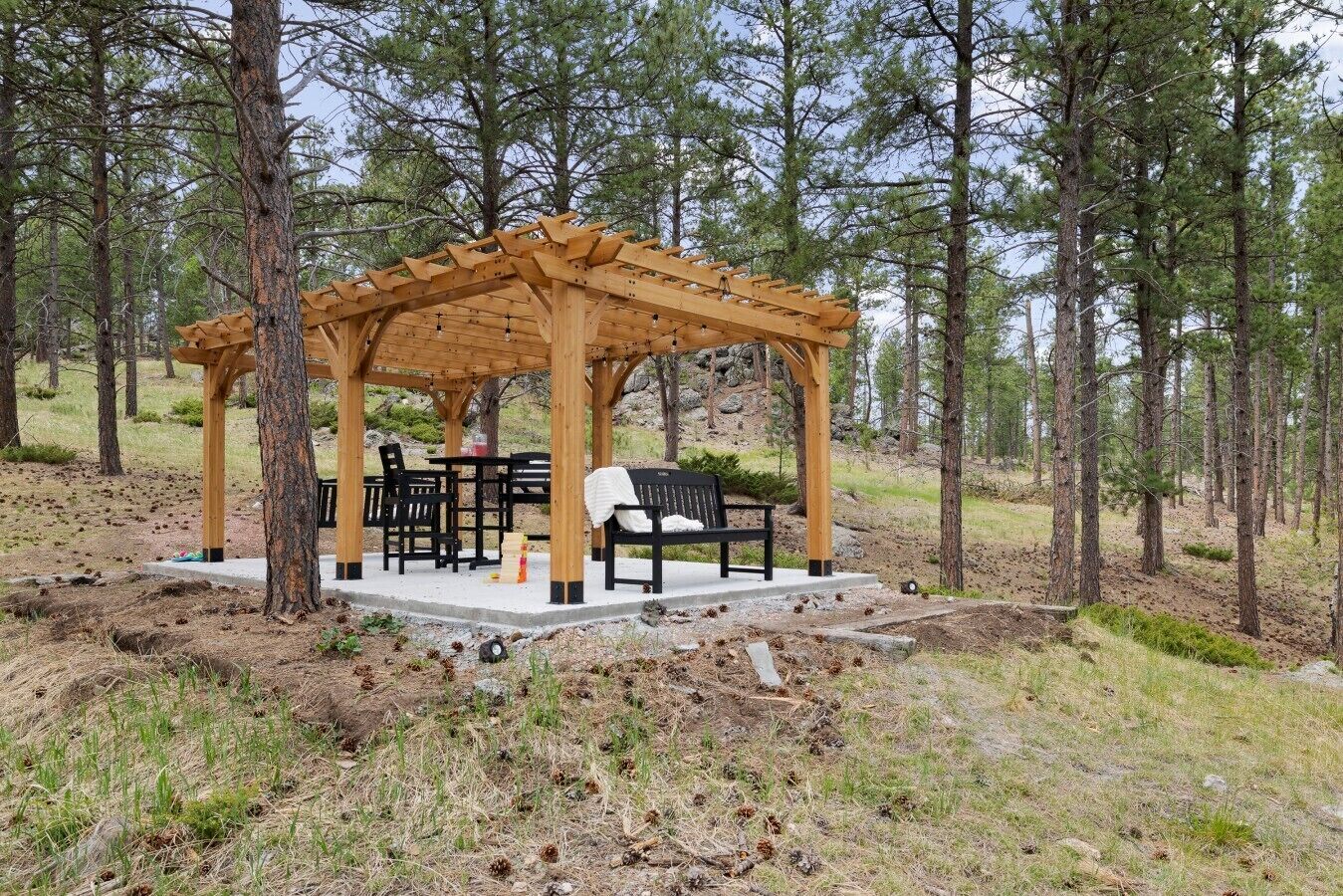 A wooden gazebo with a table and chairs, surrounded by trees.