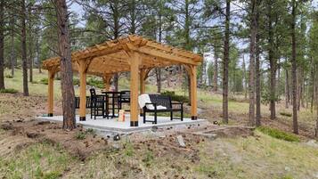A wooden gazebo with a table and chairs, surrounded by trees.