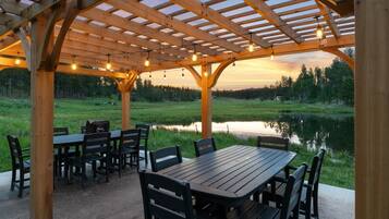 A wooden pergola with tables and chairs, overlooking a pond and forest.