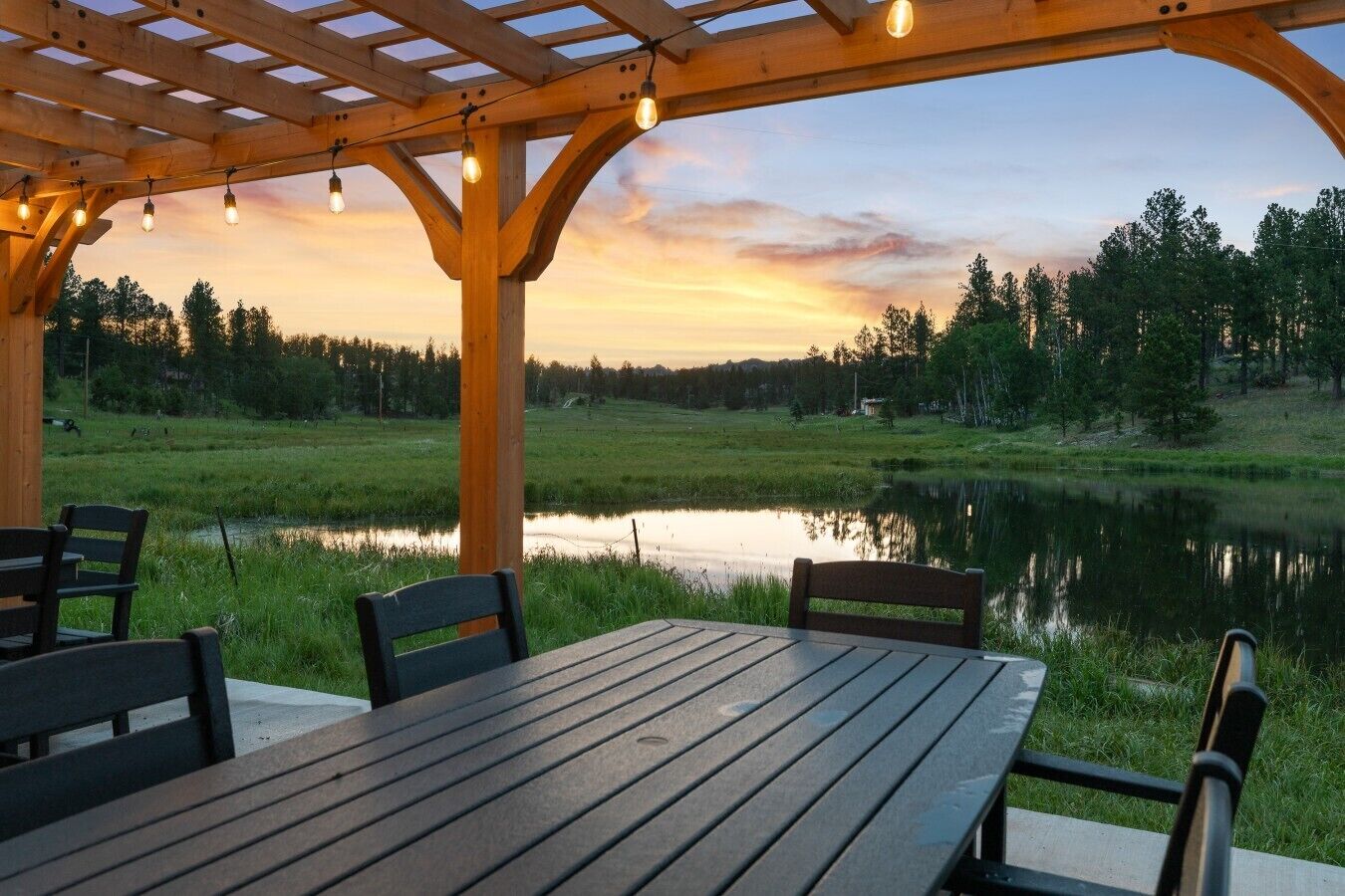 A wooden pergola with string lights, a table, and chairs overlooking a lake at sunset.
