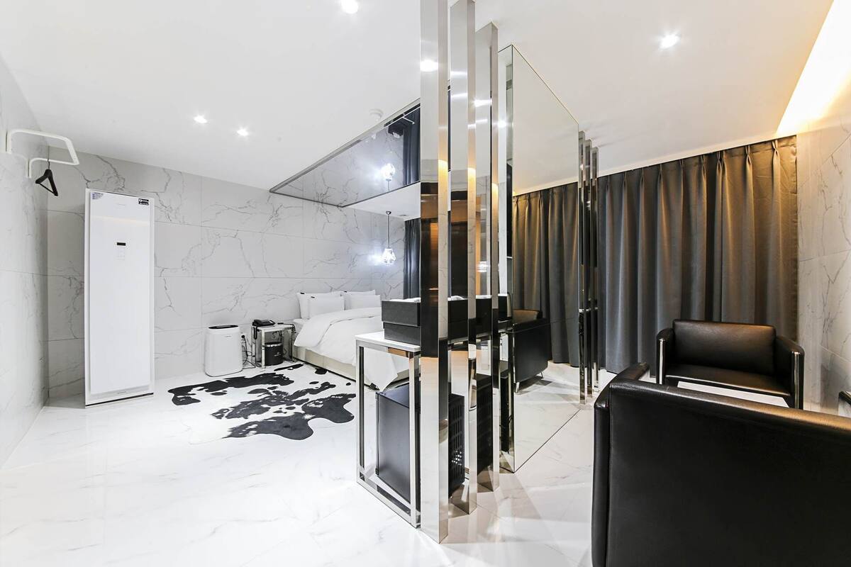 A modern hotel room with a large bed, a seating area with a black leather chair, a glass partition, and a white wardrobe.