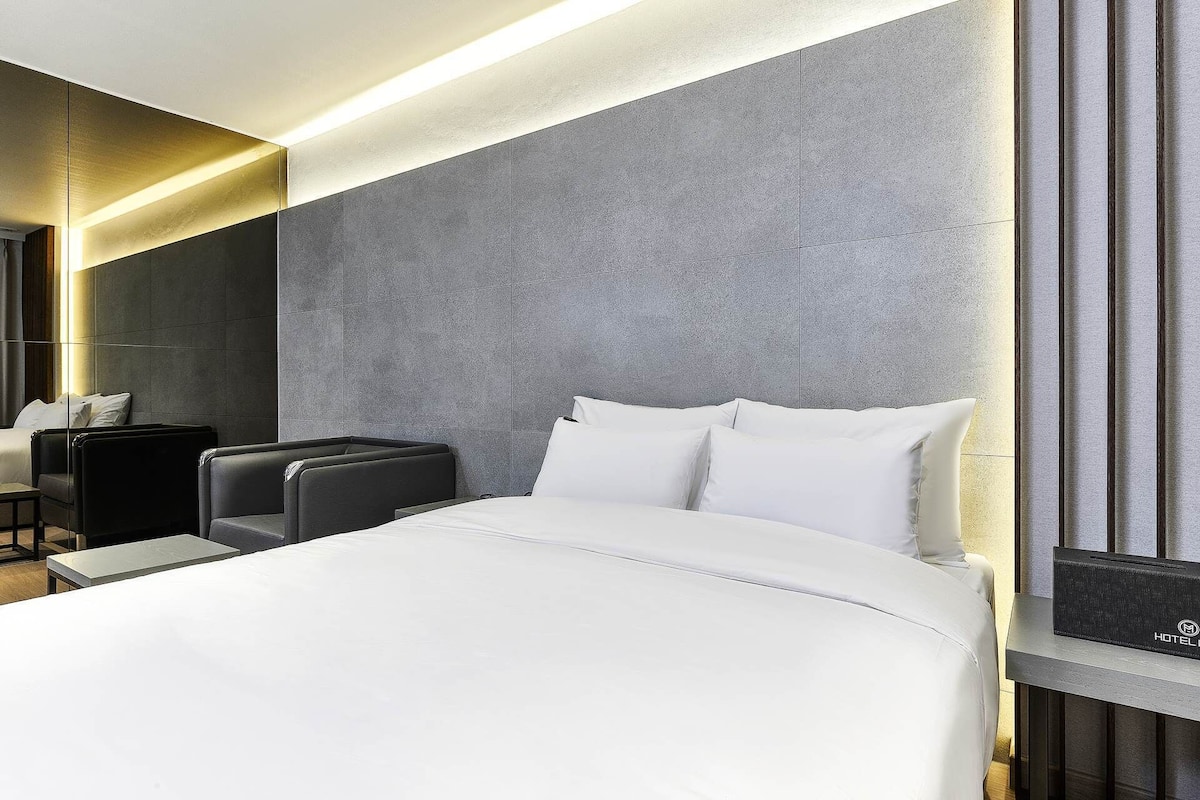A modern hotel room with a large bed, a seating area with a chair and a small table, and a large mirror on the wall.