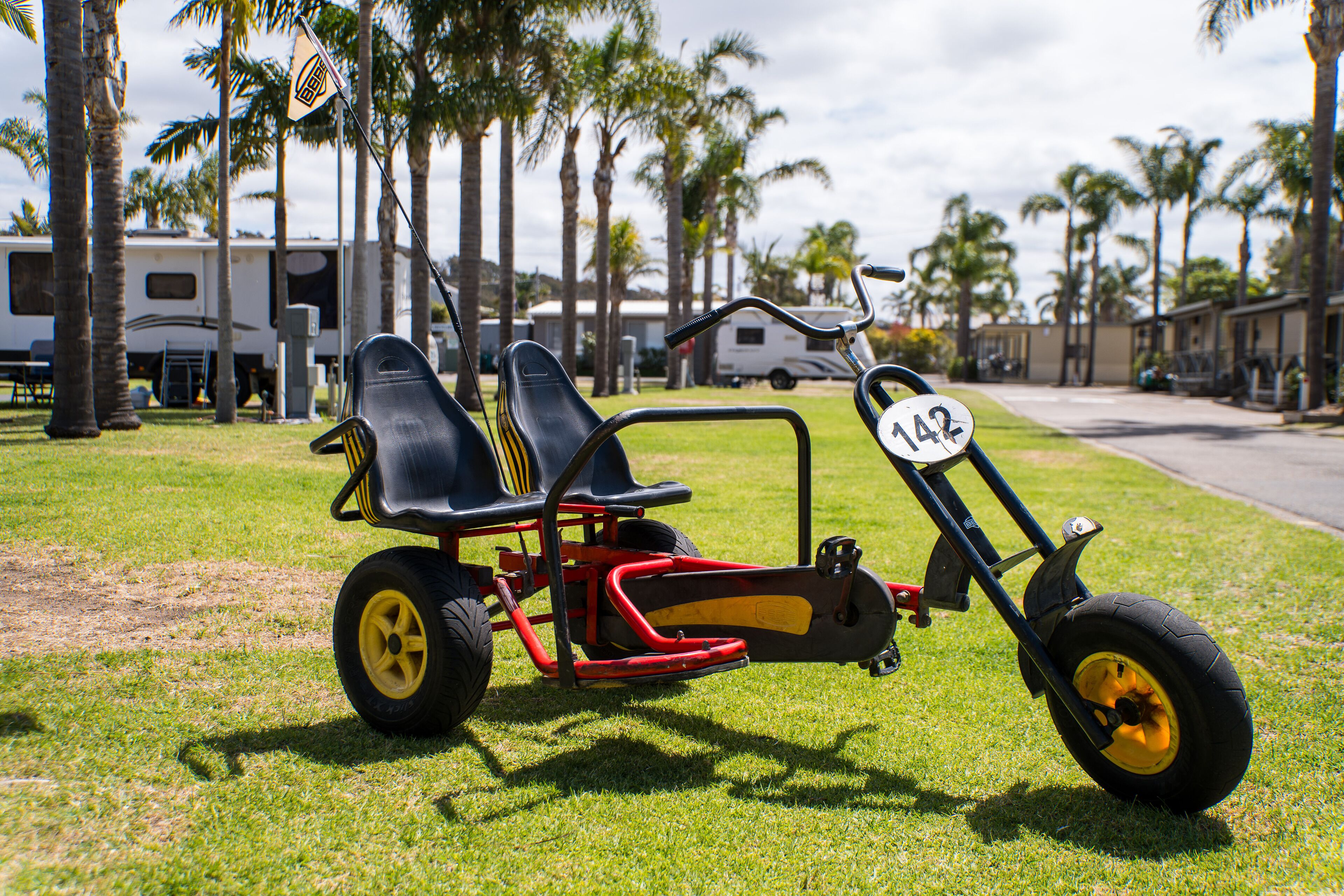 A small, three-wheeled vehicle with two seats and a number 142 on it, parked on grass with palm trees and RVs in the background.